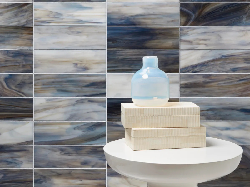 A small blue glass bottle sits on three stacked wooden boxes atop a round white table, set against a backdrop of rectangular blue and gray marbled tiles.