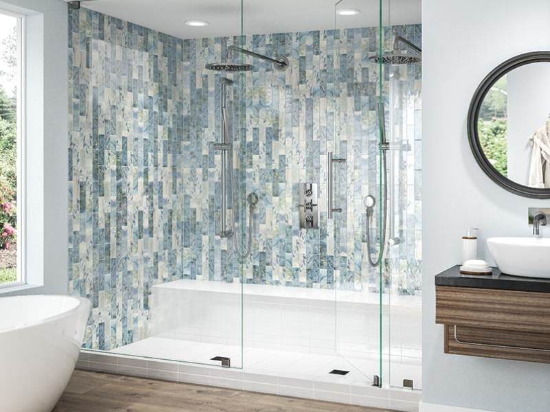 A modern bathroom features a glass-enclosed shower with blue mosaic tile walls, a double showerhead, a built-in bench, a round mirror above a floating vanity, and a white freestanding bathtub.