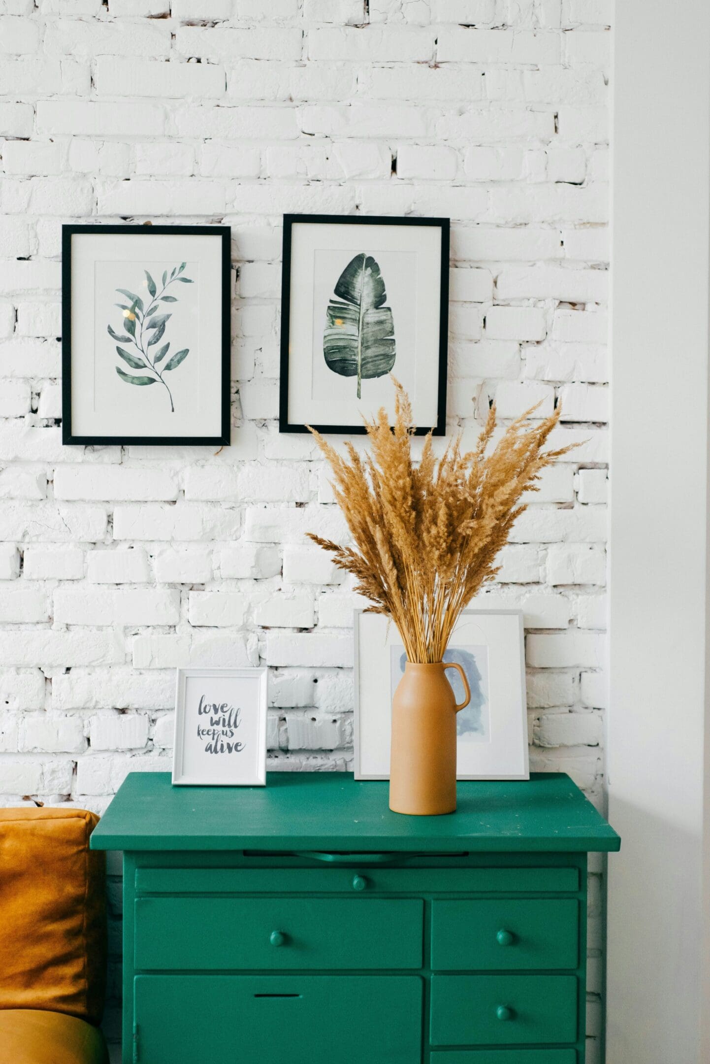 A green dresser against a white brick wall with framed botanical prints. A vase with dried grasses sits on the dresser next to a small framed quote.